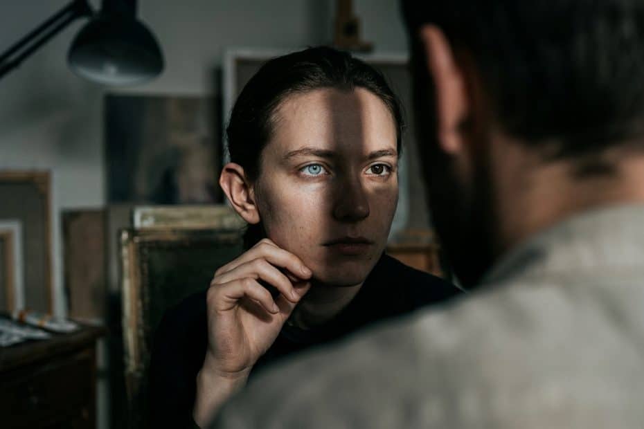 Close-up of a woman in a dim art studio, gazing at a man off-camera with her hand near her chin as a shadow crosses her face.